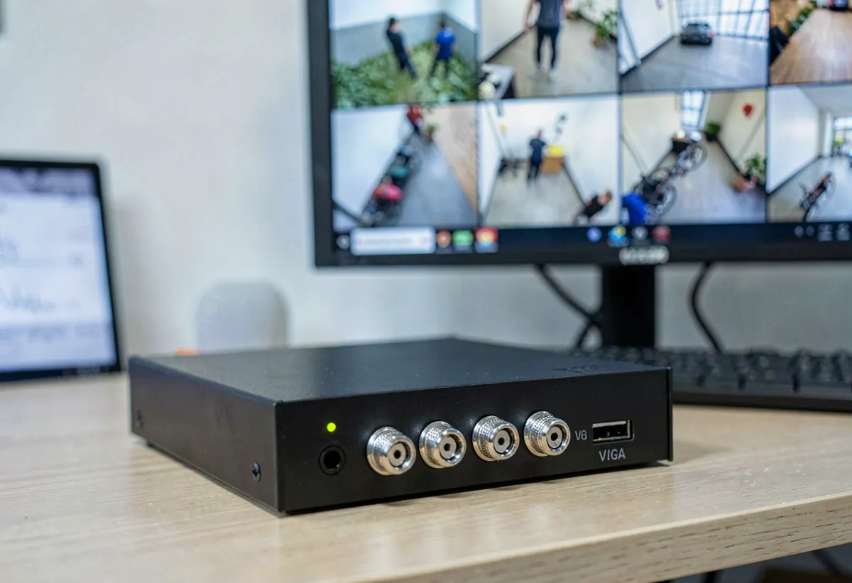 Analog security DVR with BNC connectors on a desk beside a monitor showing a multi-camera grid